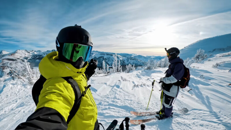 Two skiers standing on a snowy mountain peak, dressed in vibrant winter gear and helmets, with one taking a selfie against a backdrop of breathtaking snow-covered mountains