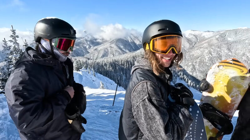 Two snowboarders, dressed in winter gear and helmets, stand on a snowy mountain slope, smiling and enjoying the clear, sunny day with a breathtaking view